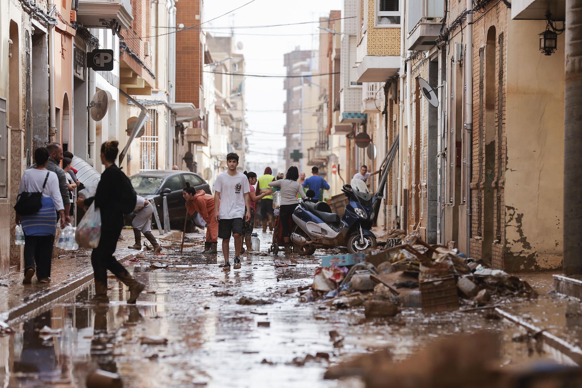 Unas personas caminan por una calle cubierta de lodo mientras otras se afanan en las labores de limpieza tras las intensas lluvias por la fuerte dana. EFE.